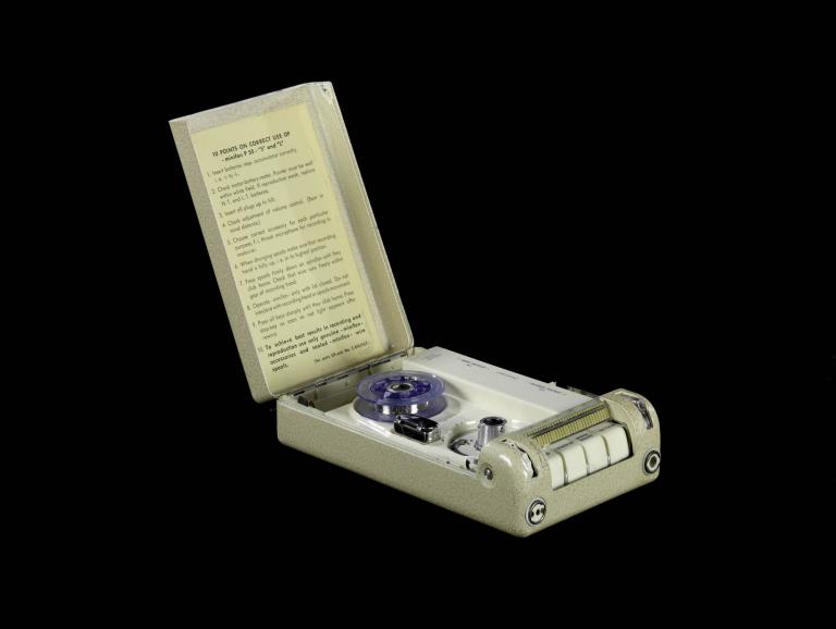 A photo of a miniature wire recorder. It is badge in colour and within the lid there's a large body of text with instructions for use.