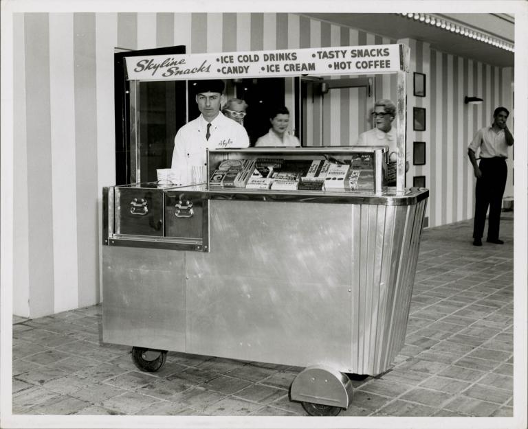 Vintage snack cart with skyline branding, offering drinks, snacks, and ice cream, outside a theater in the 1950s