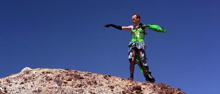 Hugo Weaving in dressed in green sequined dress dancing on a mountain in the desert