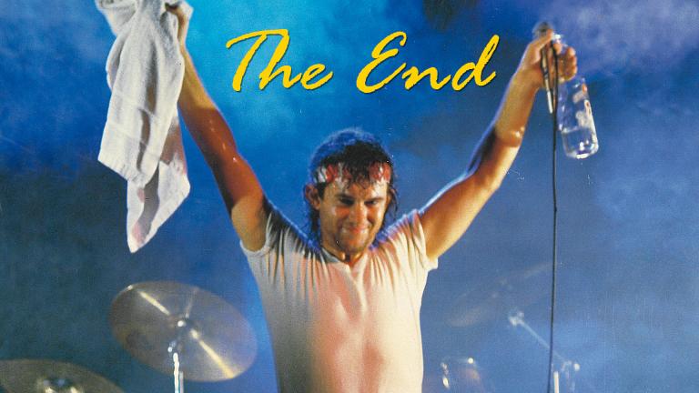 Jimmy Barnes wearing a white t-shirt on stage with his arms raised, he is holding a white towel in one hand and a microphone and vodka bottle in the other.