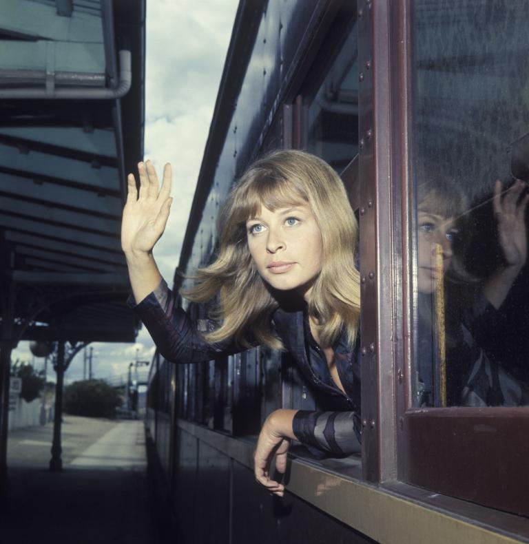 A young woman leans out the window of a train waving.