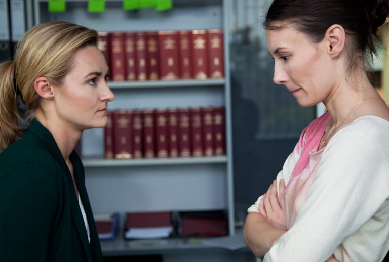 Two young women in a workplace scene pictured from waist up in profile. They are facing each other and looking confrontational, one of the women has her arms folded across her chest.