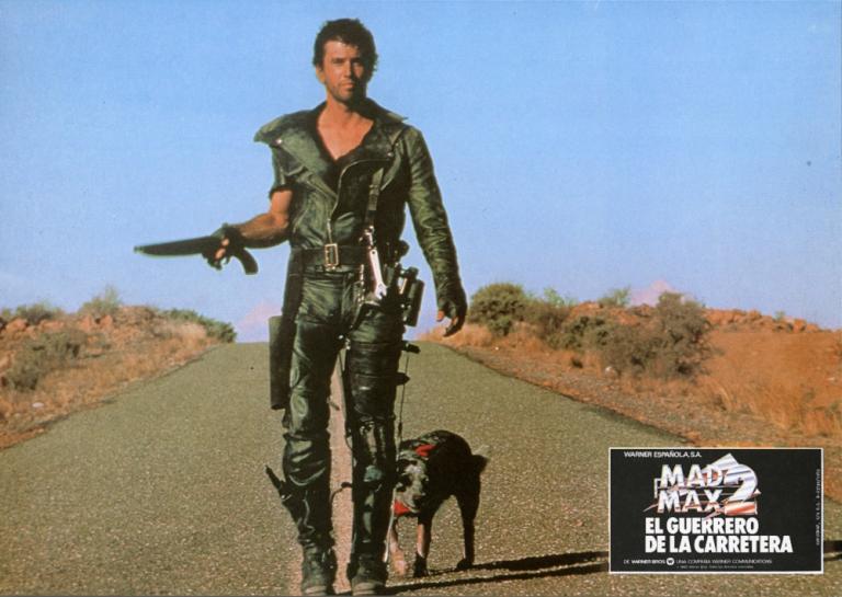 Mad Max 2 lobby card featuring Max (Mel Gibson) with his dog walking down a road holding a sawn-off shotgun.