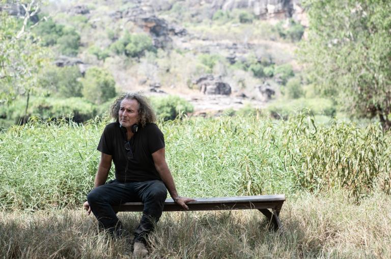 A middle-aged man in a black t-shirt and jeans sitting on a bench in a bush setting.