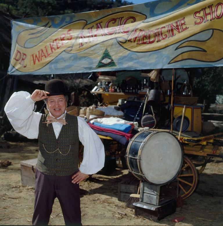 Graham Kennedy in a scene from the TV series Five Mile Creek, he is dressed as a travelling salesman in front of a sign that says Dr Walker's Magic Medicine Shop