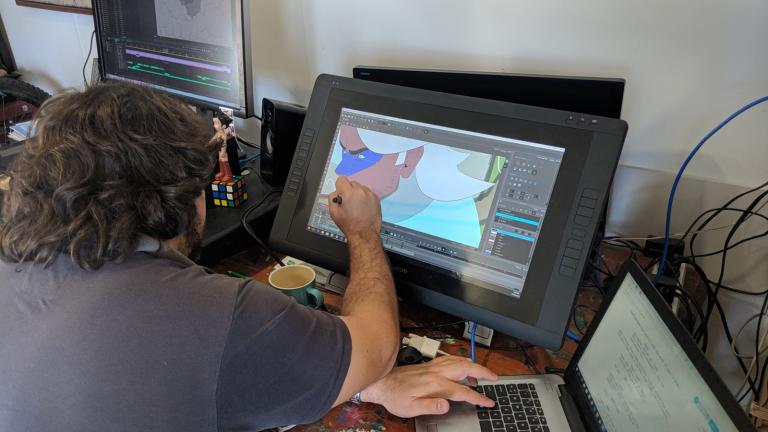 A man pictured from over his shoulder, seated in front of laptop and computer monitor. His screen shows an animated image.