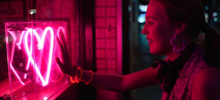 A woman places her hand against a shop window where a pink neon heart glows.