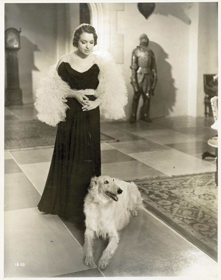 A woman wearing a formal dress stands in a large lobby. At her feet lies a  white borzoi hound.