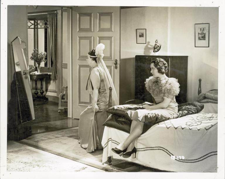 A woman wearing a ball gown and headdress looks in a long mirror while another young woman, seated on a bed, watches her.