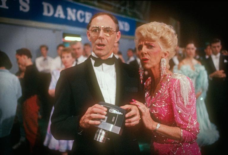 Scene from Strictly Ballroom showing an older man wearing a tuxedo and holding a video camer and an older woman in a pink sequinned dress with a concerned look on their faces. 