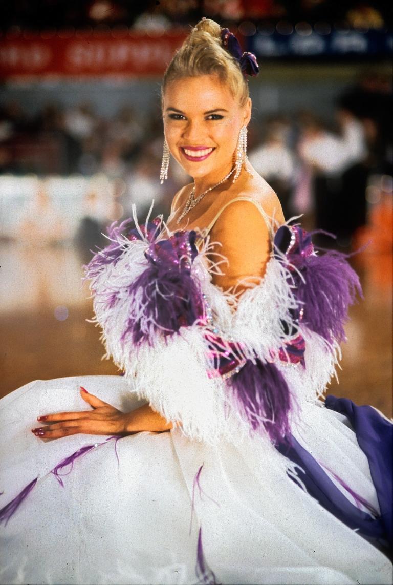 A woman smiling at camera wearing a white ballroom gown with purple feathered trim.