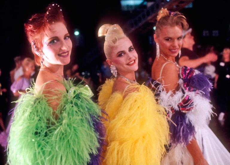Scene from Strictly Ballroom showing three women smiling at the camera wearing colourful ballroom gowns with feather trim.