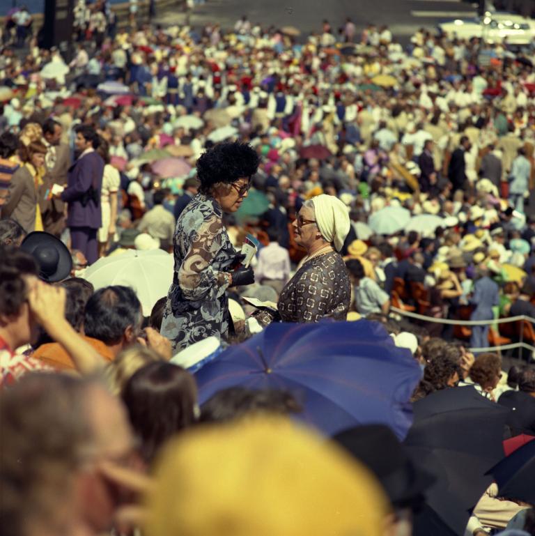 Two women dressed in dresses and hats talk to each other. They are surrounded by a massive crowd of people.