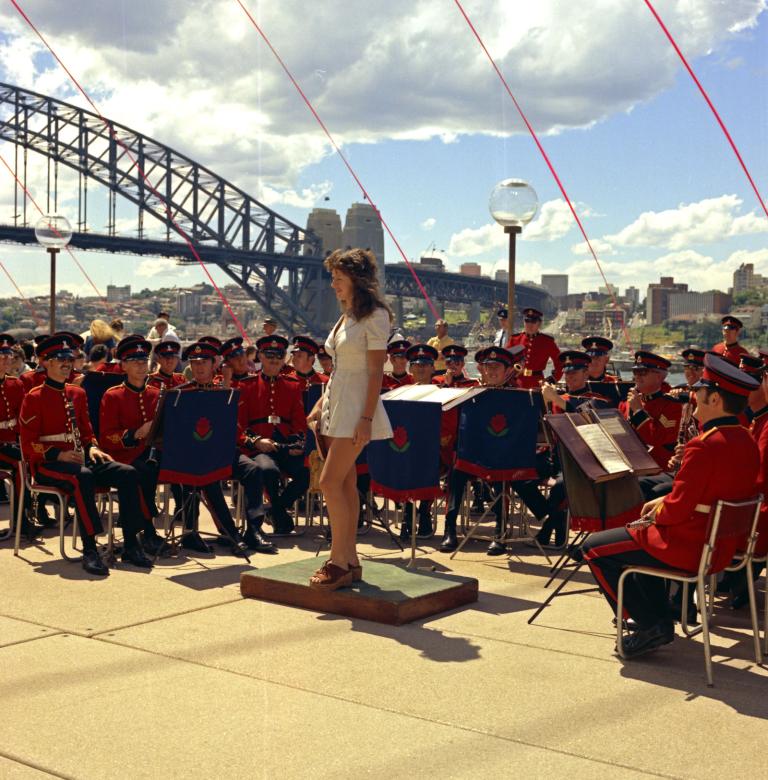 A woman in a white minidress stands, surrounded by a military band. The Sydney Harbour Bridge is in the background.