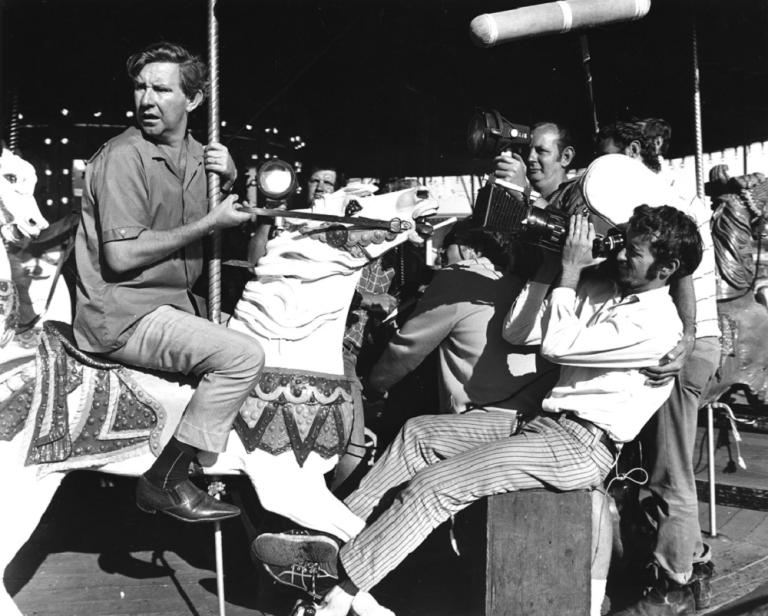 John Meillion being filmed riding a horse on a merry-go-round.