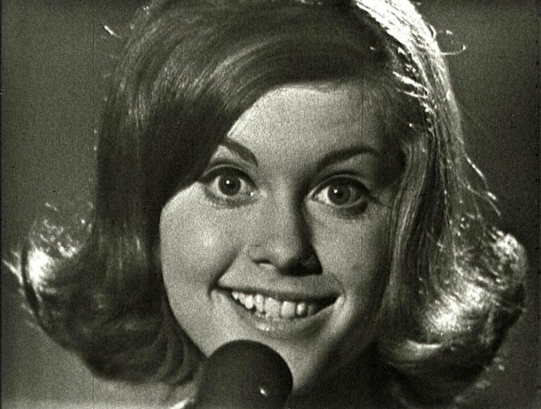 Close up of Olivia Newton-John smiling and looking directly at the camera with a microphone in front of her face, partially covering her chin, from an episode of 'Boomeride' when she was aged 16.