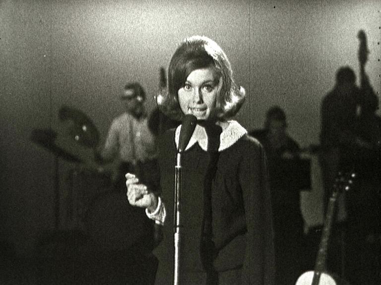 Olivia Newton-John at age 16 in a TV studio standing behind a microphone singing.