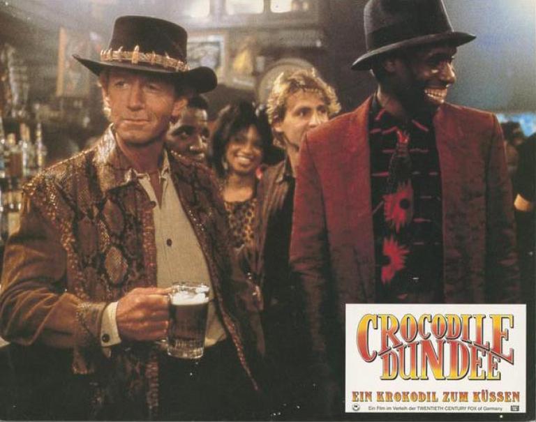 Lobby card depicting Mick Dundee standing in a bar with a beer in his hand and a guy smiling next to him like Mick's just said something funny