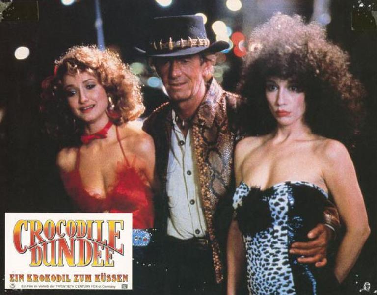 Lobby card depicting Mick Dundee with his arms around two New York City prostitutes