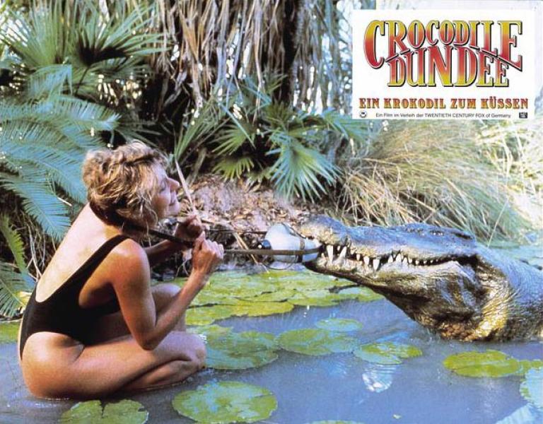German lobby card for Crocodile Dundee depicting Sue Charlton wrestling to get a water bottle off from around her neck because a crocodile has got a hold of it 