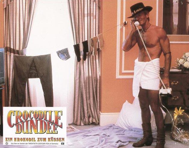 Doing the laundry outback style Lobby card depicting Mick Dundee talking on the phone in his hotel room while he stands wrapped in a towel. He has made a makeshift laundry for his clothes across the window.