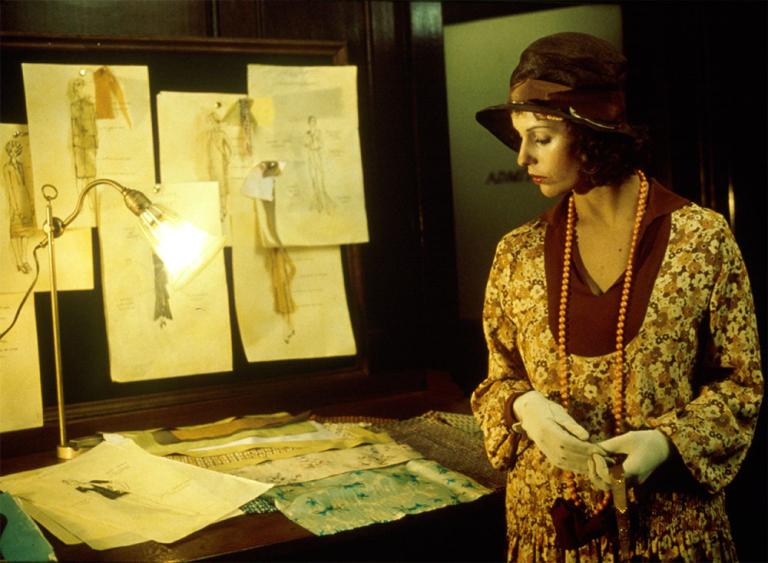 Film still of Helen Morse as Caddie looking at drawings of women's dress designs in a studio.