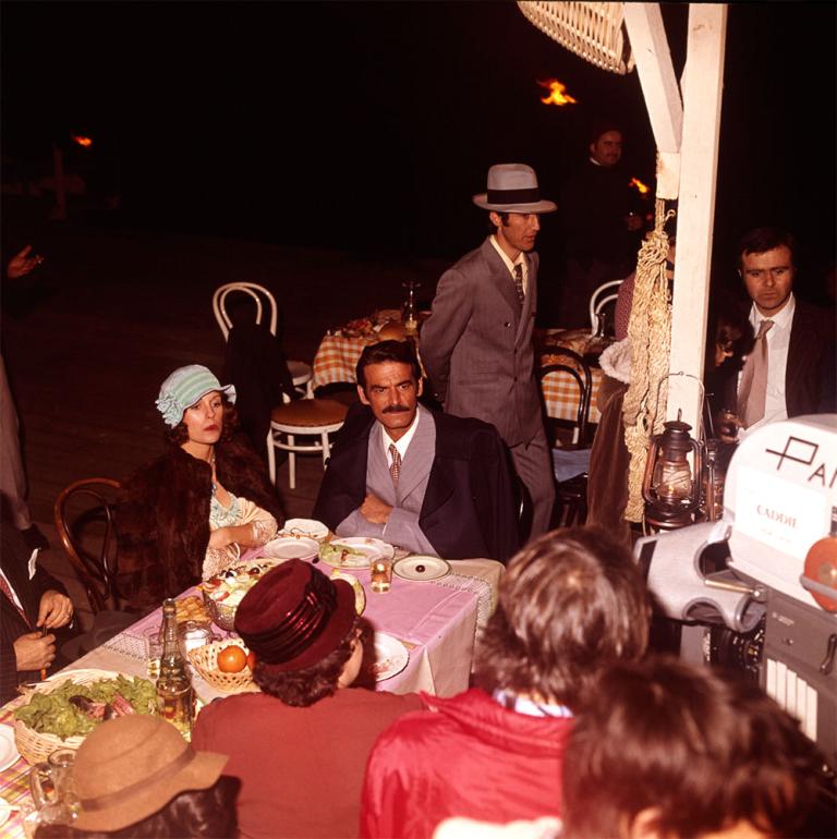 Behind the senes shot of Helen Morse as Caddie and Takis Emmanuel as Peter on the set of Caddie. They sit at a outdoor dining table while the camera crew and extras work around them.
