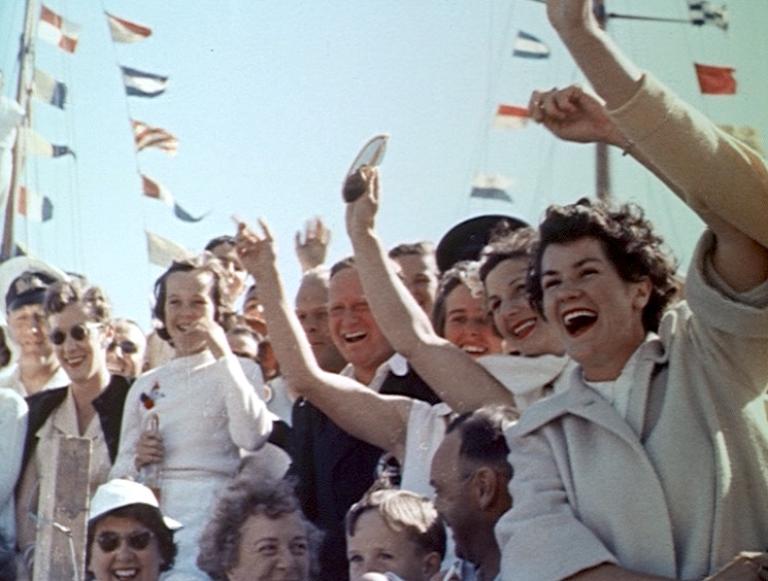 A crowd of people wave enthusiastically at Queen Elizabeth II on her visit to Hobart in 1954. There are flags in the background.
