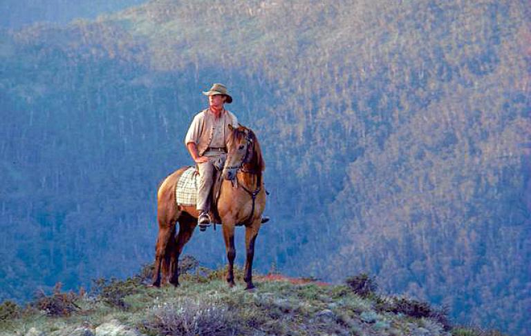 Jim Craig (played by Tom Burlinson) sits in the saddle on a horse on the top of a hill with rugged mountains in the background, in a scene from 'The Man From Snowy River'