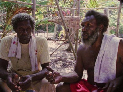 Eddie Mabo is seated on the ground, talking with another man.
