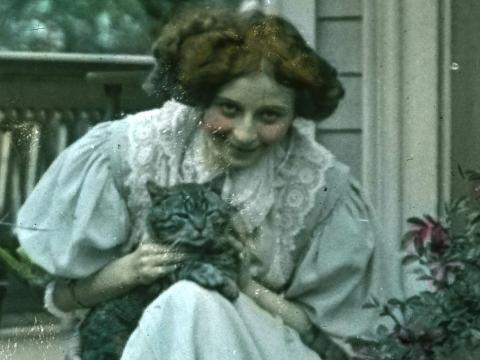 Young woman sitting on some steps in front of a house and holding a grey cat, circa 1906.