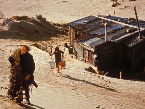 Peter Cummins as Hideaway Tom walking up the sand dunes with fishing nets slung over his shoulder and Greg Rowe as Storm Boy following him the dunes carrying two pails of water. Their simple corrugated iron humpy is below them.