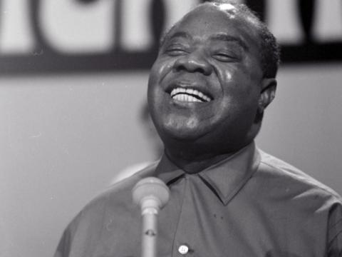 Close up, head and shoulders shot of jazz musician Louis Armstrong singing into a microphone with his eyes closed, circa 1964.