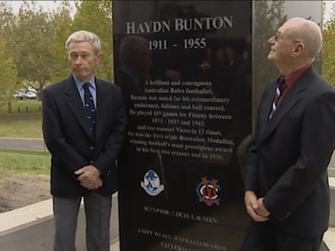Two older men wearing suits standing either side of the base of a statue.