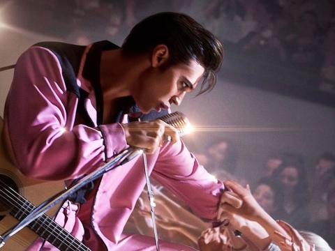 Actor Austin Butler in a scene from the film ELVIS. He is on stage wearing a bright pink suit and leaning out towards the crowd.