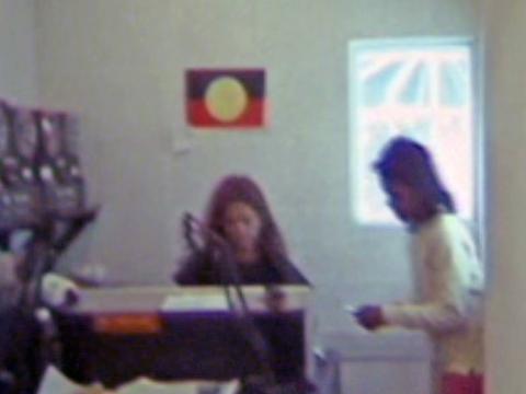 Two people in a radio studio. There is an aboriginal flag on the wall behind them.