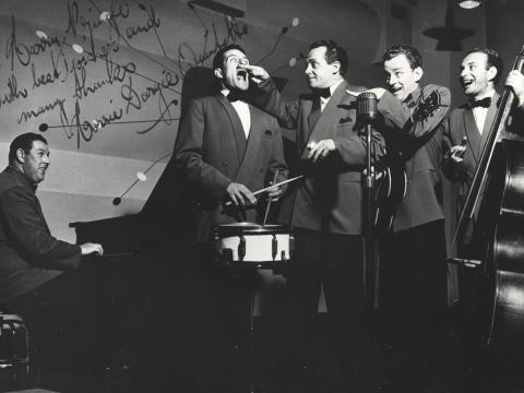 A group of five men in a band. One is sitting down at a piano and the others are standing.
