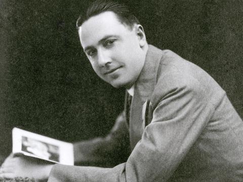A man in a suit pictured from the waist up in profile leaning forward with his head turned towards the camera with a book open in front of him.