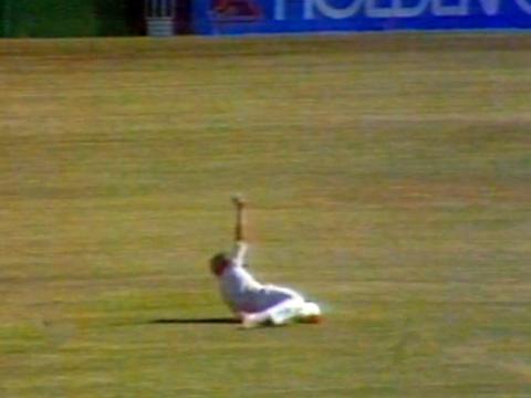 A man lying on his side on a cricket oval holding his arm straight up. He is holding a cricket ball in his hand.