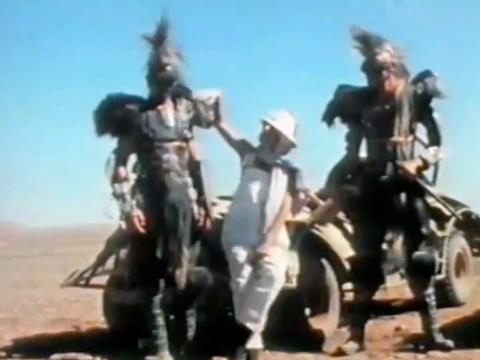Two men wearing futuristic costumes standing either side of a woman dressed in white clothes and hat.