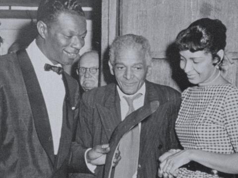 Singer Nat King Cole holding a boomerang and standing alongside an older First Nations man, William Bodney, at the Coolbaroo Club, c1955. Cole's wife Maria is standing to the right. 