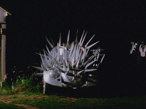 A silver Volkswagen covered in spikes like an echidna, and parked on a country road at night