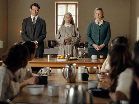 A man and a woman standing either side of a nun, addressing a room full of young First Nations schoolgirls.