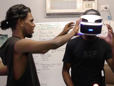 Two young First Nations men standing in an office. One is placing a VR helmet on the other's head.