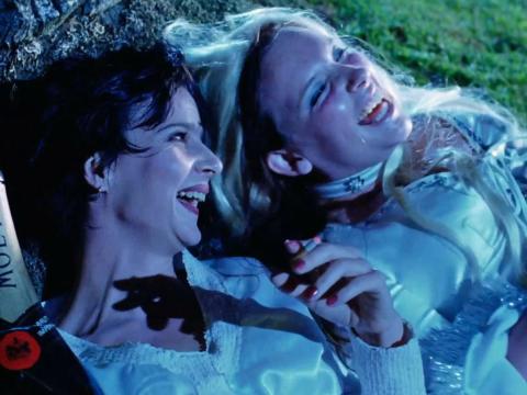 Rachel Griffiths and Toni Collette lying side by side on the grass laughing in a scene from the film Muriel's Wedding. Griffiths is clutching a bottle of champagne and smoking a cigarette