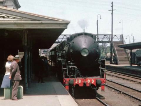 A steam locomotive pulling into a train station. A man and woman stand on the railway platform with their suitcase waiting to board.