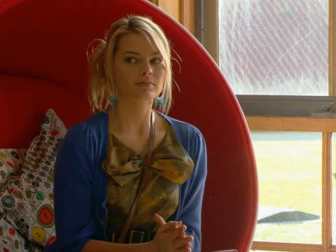Margot Robbie wearing a blue cardigan and seated on a large chair by a window