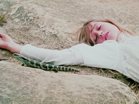 Miranda lies asleep on a rock, arm outstretched, in a scene from Picnic at Hanging Rock. A large lizard passes by her immobile form. 