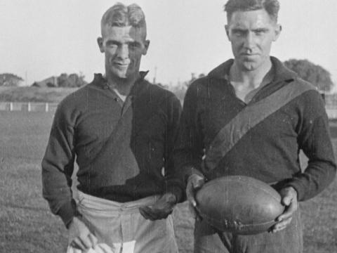 Two 1920s AFL players standing on the pitch in uniform. They are facing the camera and one is holding a football