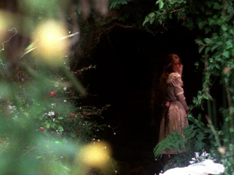 Miranda stands in a darkened hollow in a forest and looks back over her shoulder smiling.
